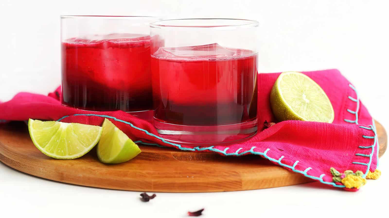 Two glasses of red hibiscus tea on a wooden platter, surrounded by lime wedges and a vibrant pink cloth. The setup is bright and inviting, with a fresh, colorful presentation.