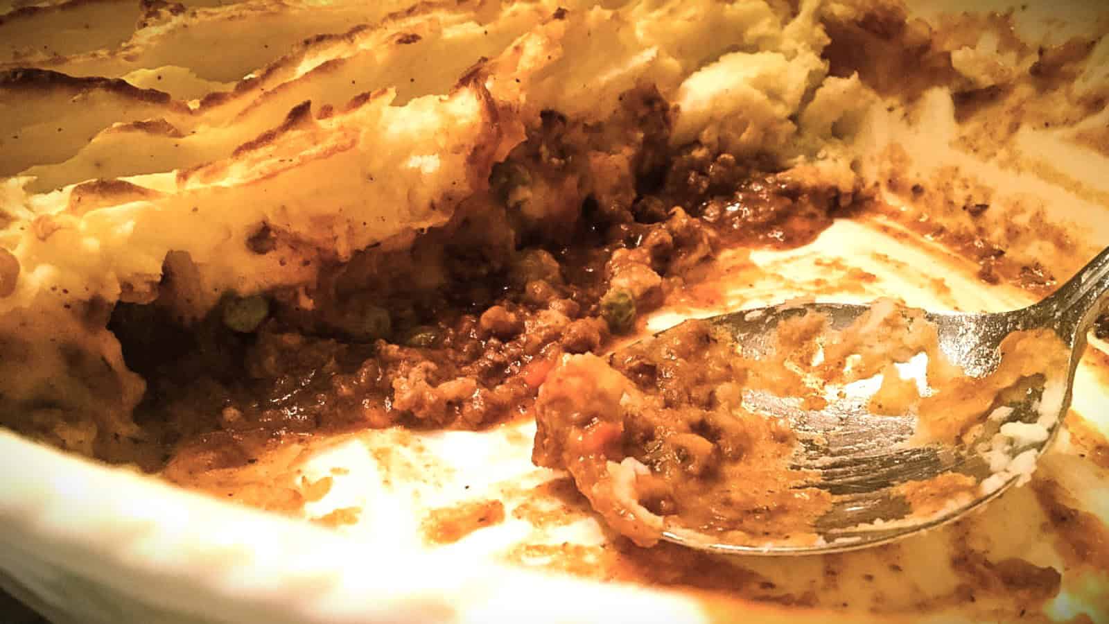 A close-up of a casserole dish with a half-eaten shepherd's pie. The remaining portion shows layers of mashed potatoes on top of a savory meat and vegetable mixture. A spoon rests in the dish, slightly smeared with the filling.