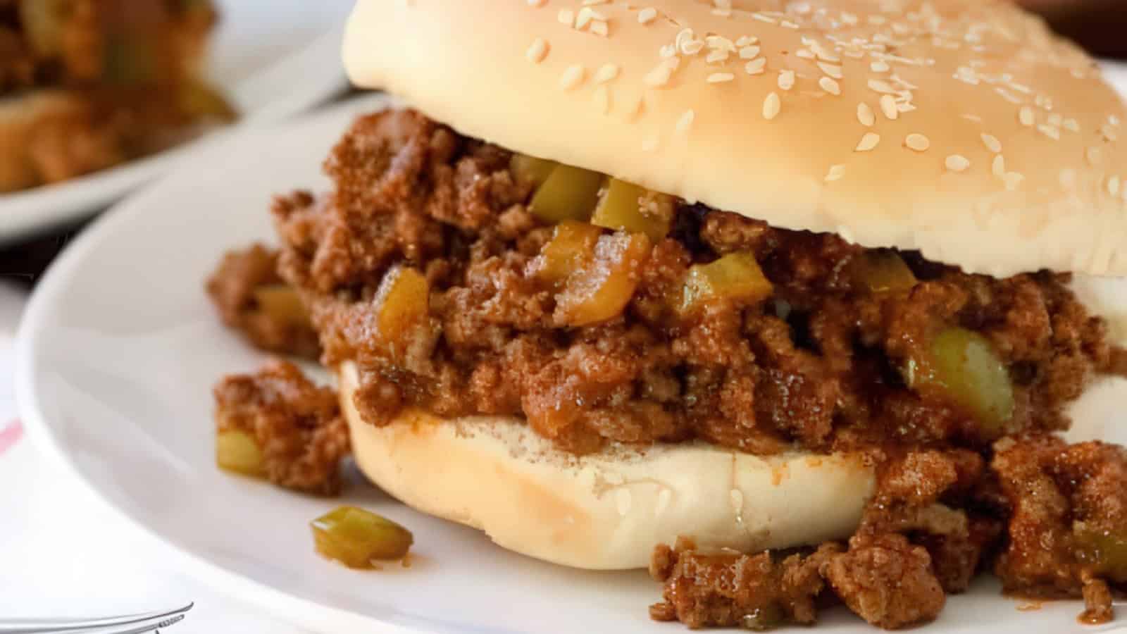 A close-up of a sloppy joe sandwich on a white plate. The sandwich consists of seasoned ground meat mixed with diced green peppers, spilling out from a soft sesame seed bun.