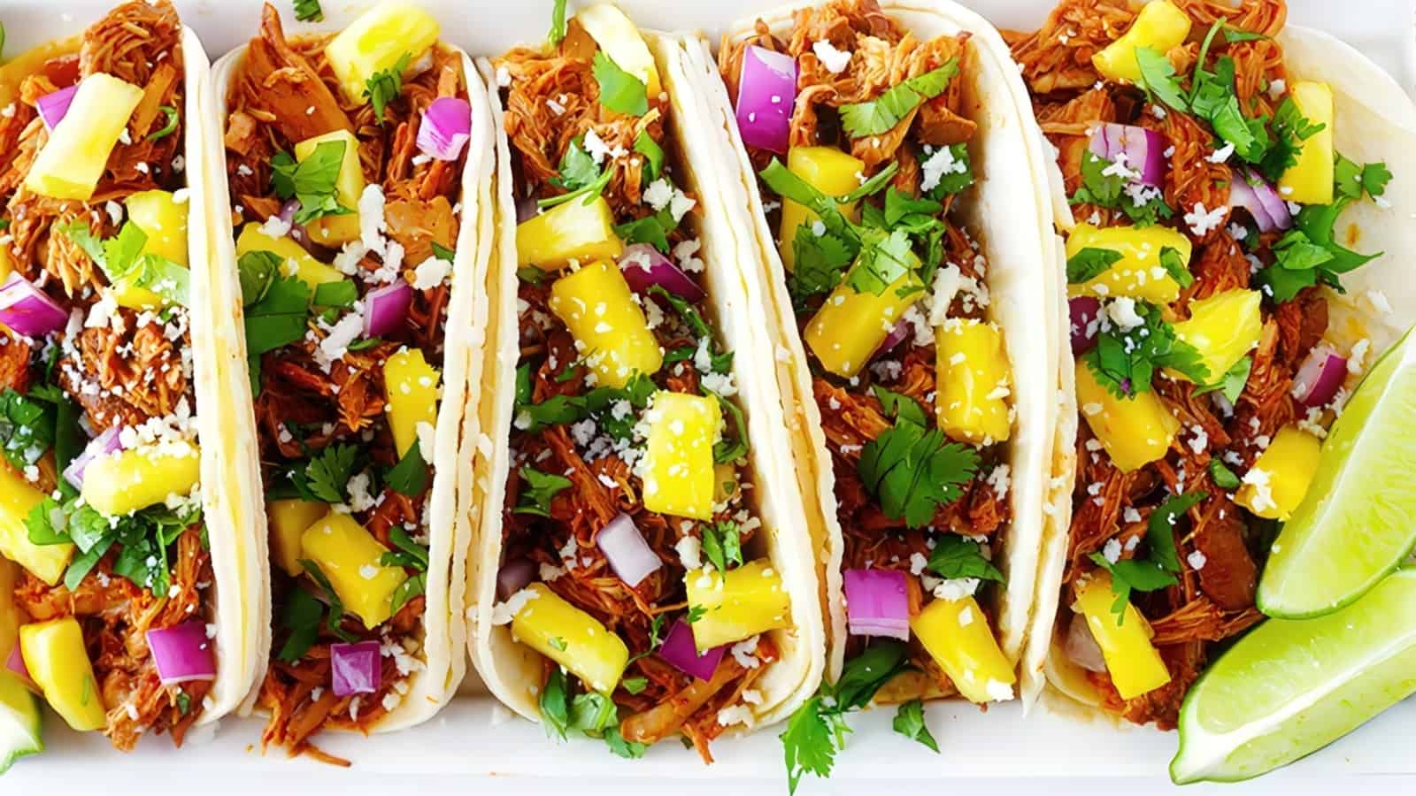 Five tacos filled with shredded meat, diced red onions, pineapple chunks, fresh cilantro, and crumbled cheese, served on a white plate with lime wedges on the side.