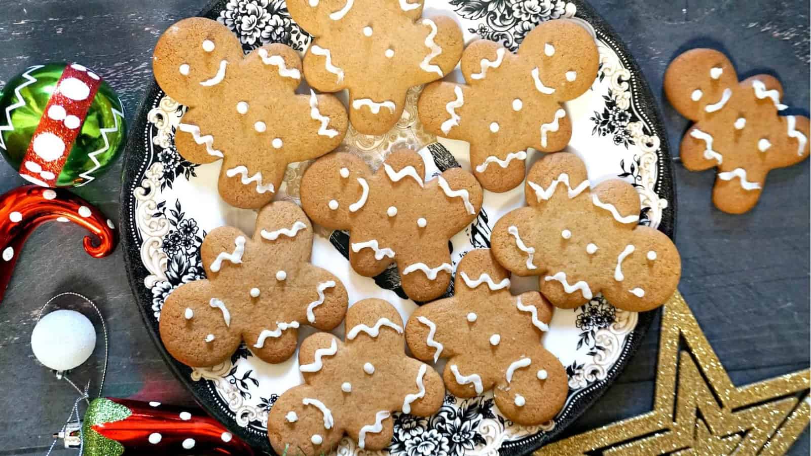 A decorative plate filled with gingerbread cookies, shaped like people and decorated with white icing, sits on a table. Festive ornaments, including a striped bauble and a glittery star, are scattered around.