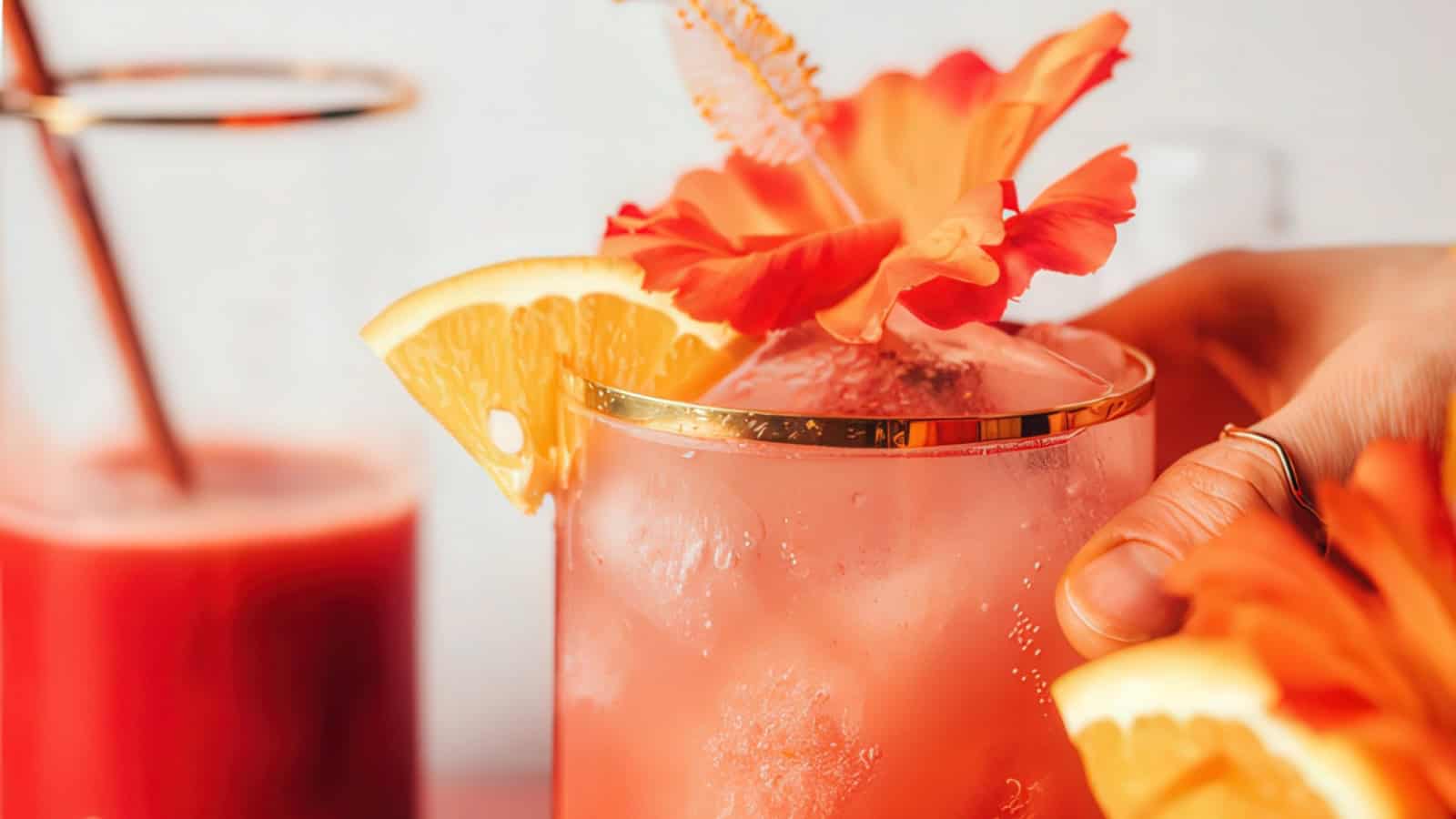 A close-up of a refreshing pink cocktail in a glass with a gold rim. The drink is garnished with a vibrant orange slice and a decorative orange flower. Another glass of red liquid is blurred in the background. A hand is holding the cocktail glass.