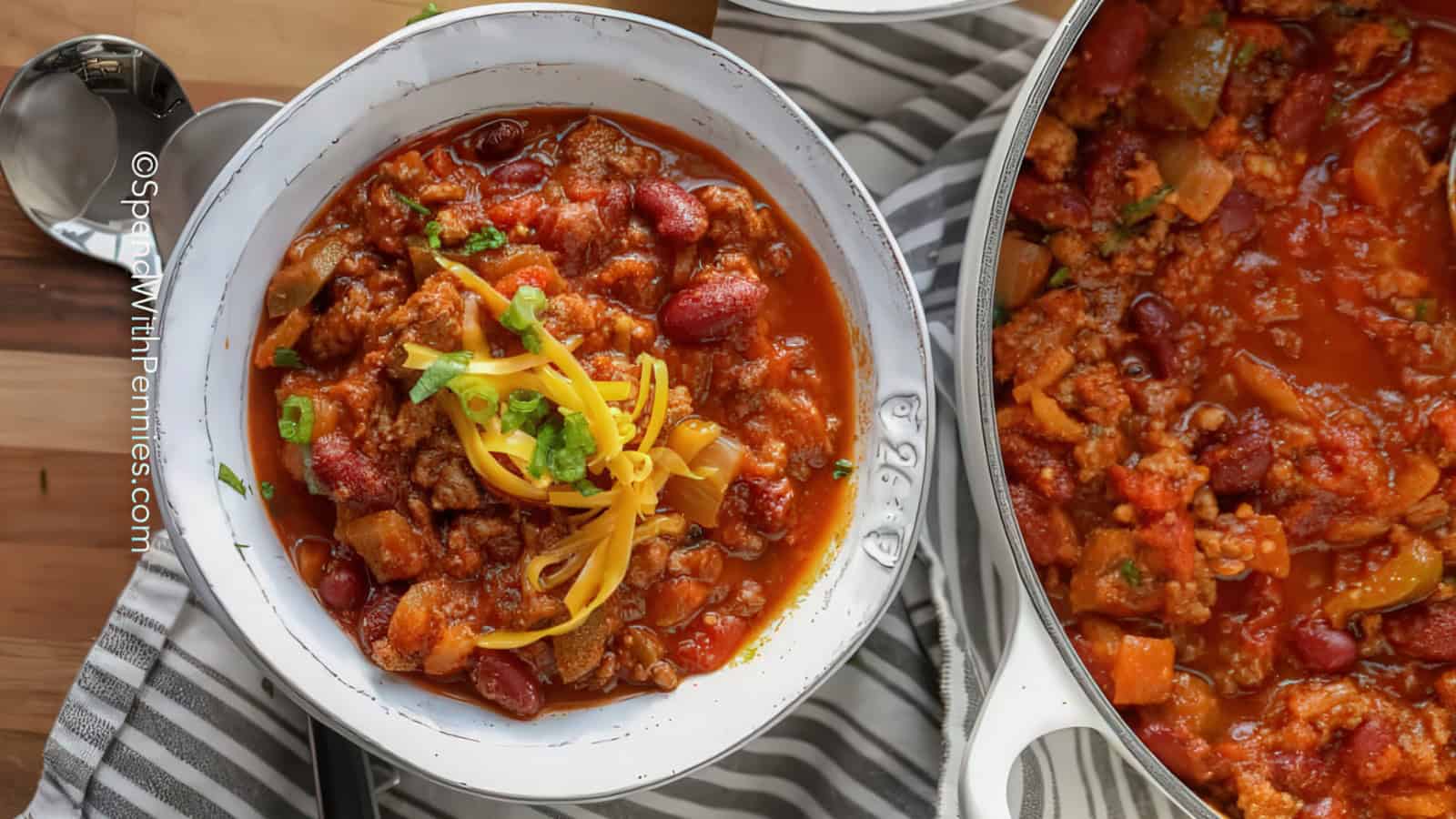 A bowl of hearty chili topped with shredded cheese and green onions next to a pot filled with more chili. The dish is rich with ground meat, beans, and vegetables, all set on a striped cloth with a spoon nearby.