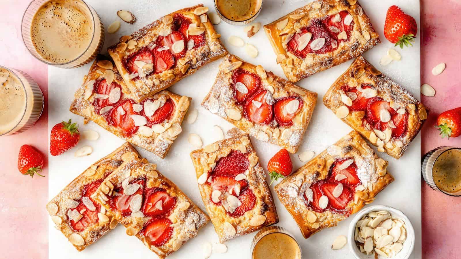 Assorted strawberry almond pastries laid out on a surface with fresh strawberries, almond slices, and cups of coffee. The pastries have a golden crust topped with sliced strawberries and almonds, dusted with powdered sugar.