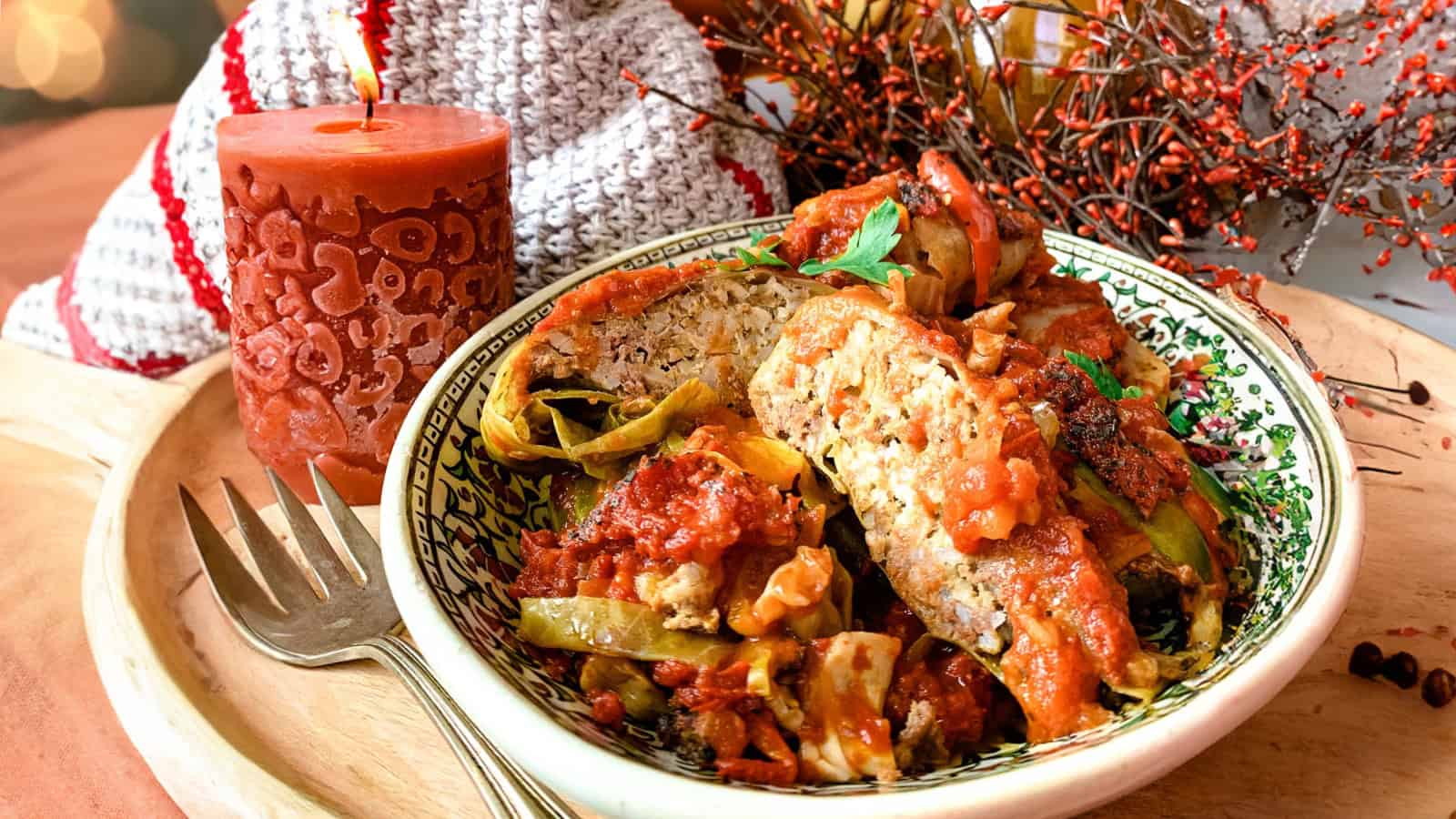 A bowl of stuffed cabbage rolls topped with tomato sauce is placed next to a fork and a lit orange candle on a decorated table.