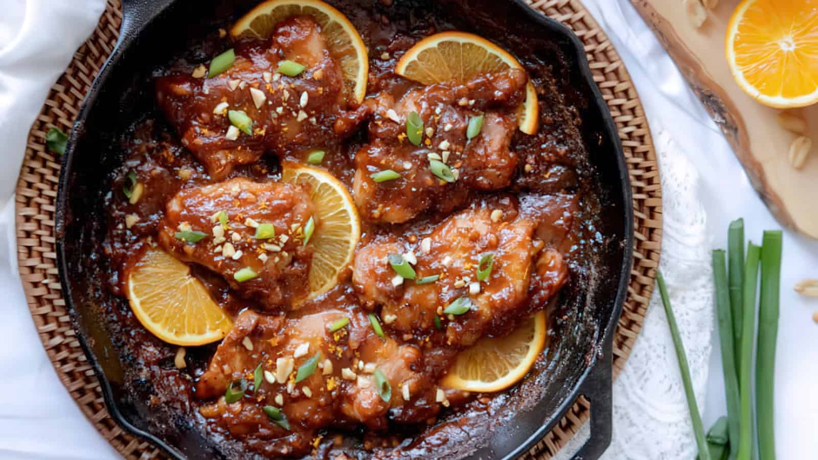 A skillet of glazed chicken thighs garnished with orange slices, green onions, and sesame seeds. The dish is set on a woven mat, with a halved orange, green onions, and white fabric in the background.