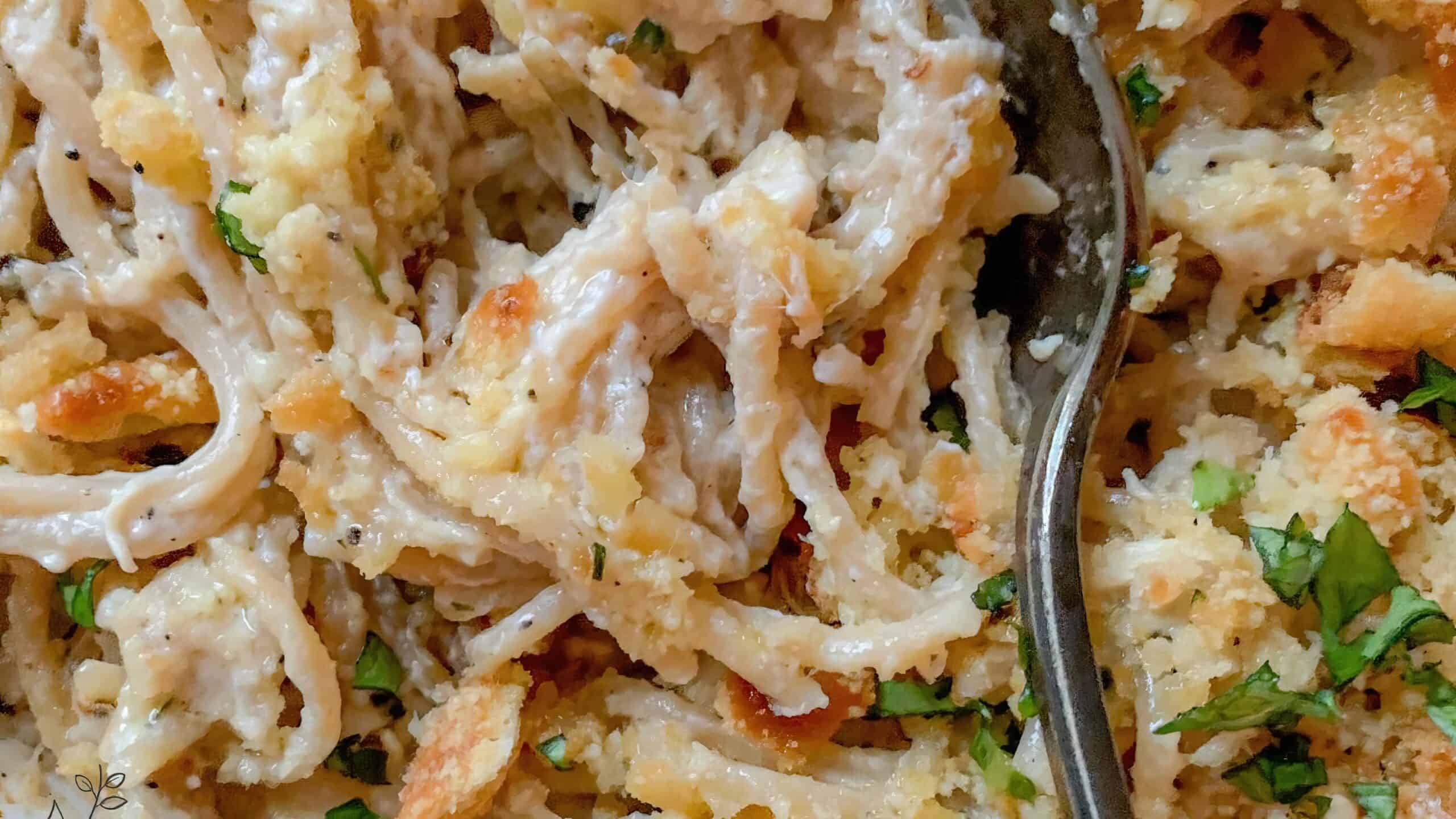 Close-up of a creamy, pasta dish topped with breadcrumbs and chopped herbs. A spoon is partially visible, mixing the ingredients. The texture looks rich and savory, with specks of green adding a fresh contrast.