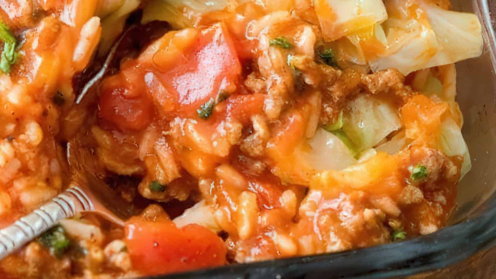 Close-up of a casserole dish containing a mix of chopped tomatoes, ground meat, shredded cabbage, and melted cheese. The ingredients are mixed together, creating a hearty and colorful texture. A spoon is partially visible on the side.
