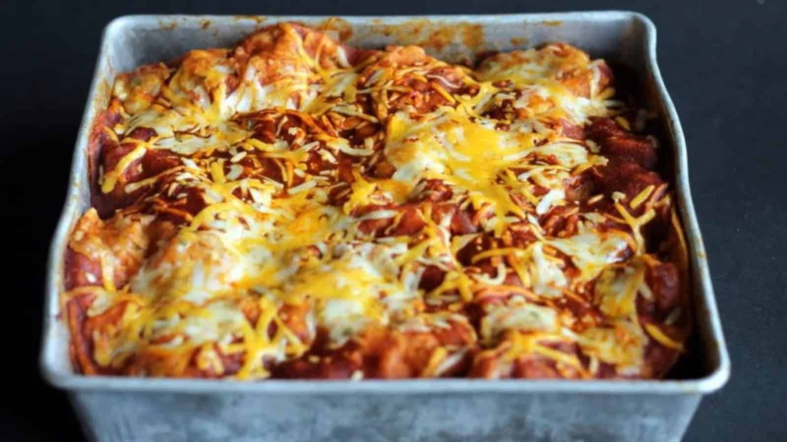 A square baking pan filled with golden-brown enchiladas covered in melted cheese and sauce. The dish has a slightly crispy texture on top, with a mix of different cheese colors, creating an appetizing, hearty look.