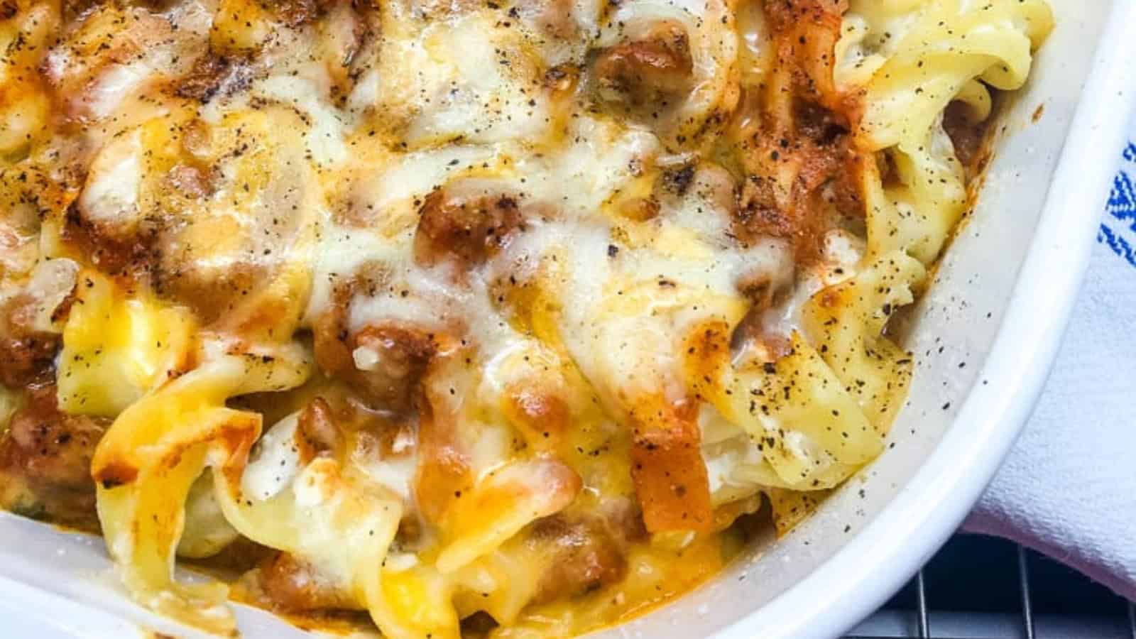 A close-up of a baked casserole dish with bubbling cheese and pepper on top, containing pasta and browned meat sauce. The edges of the casserole are golden brown, suggesting it's freshly baked.