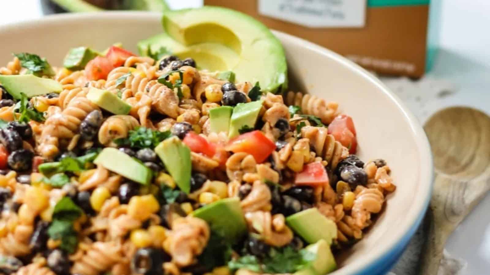 A vibrant pasta salad in a bowl, featuring rotini, black beans, corn, diced avocado, tomato, and fresh cilantro. A sliced avocado rests on the rim. A wooden spoon and a carton are in the background.