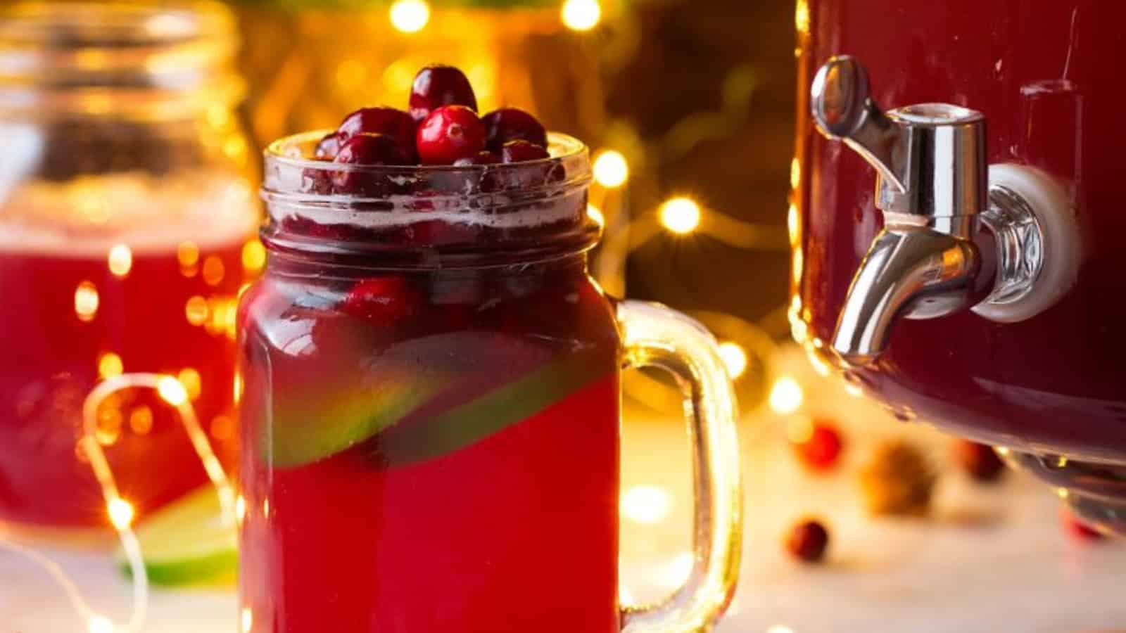 A mason jar filled with a red beverage topped with cranberries and lime slices sits on a table. Next to it is a drink dispenser with a spout. Warm, glowing string lights create a festive atmosphere in the background.