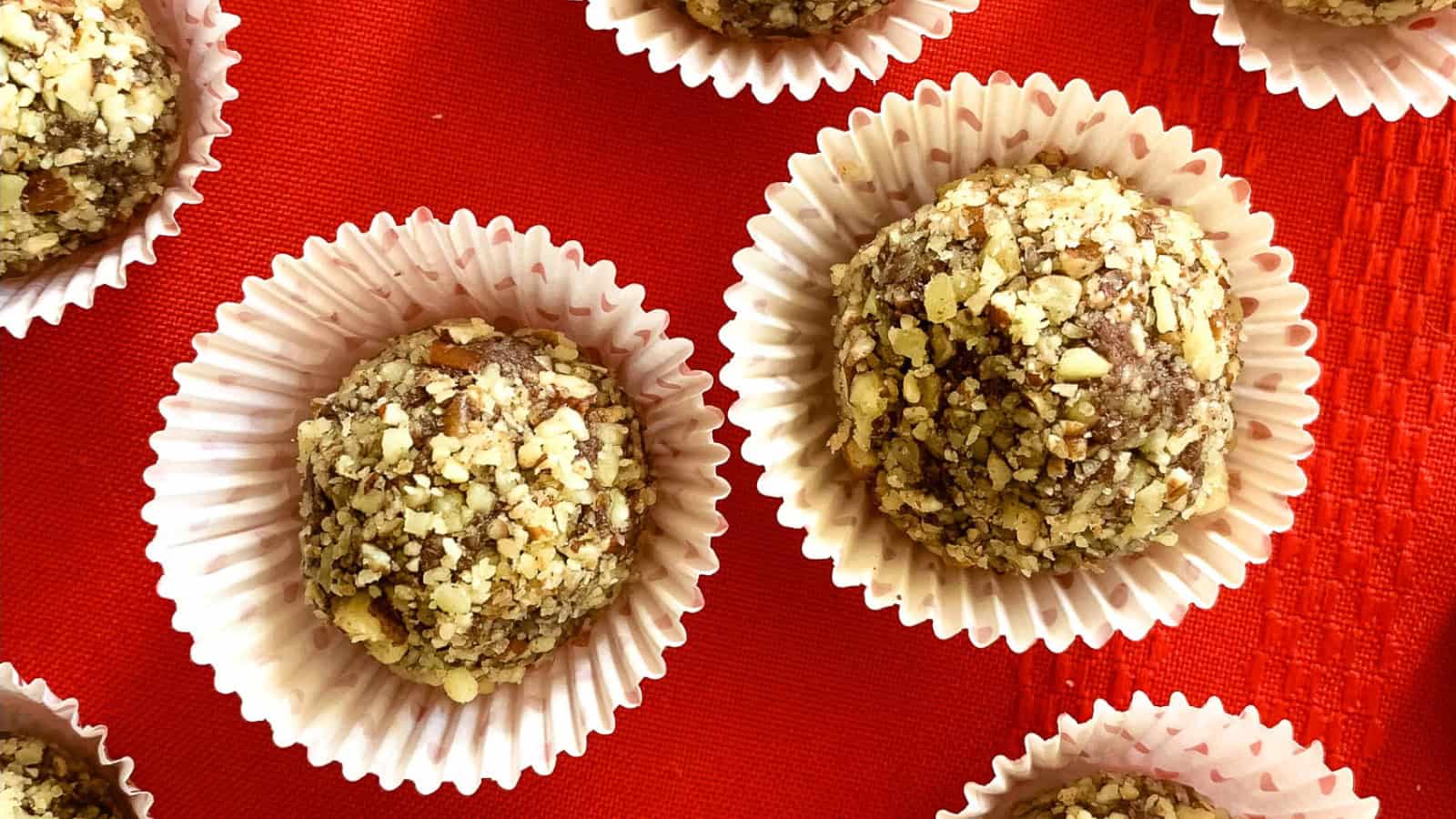 Close-up of several round chocolate truffles coated in crushed nuts, each nestled in individual white paper cups, arranged on a vibrant red surface.