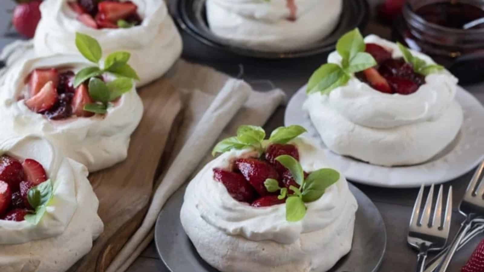 Plates with pavlovas topped with fresh strawberries and green mint leaves are arranged on a wooden board. Silver forks and a jar of jam are placed nearby, creating an elegant dessert setting.