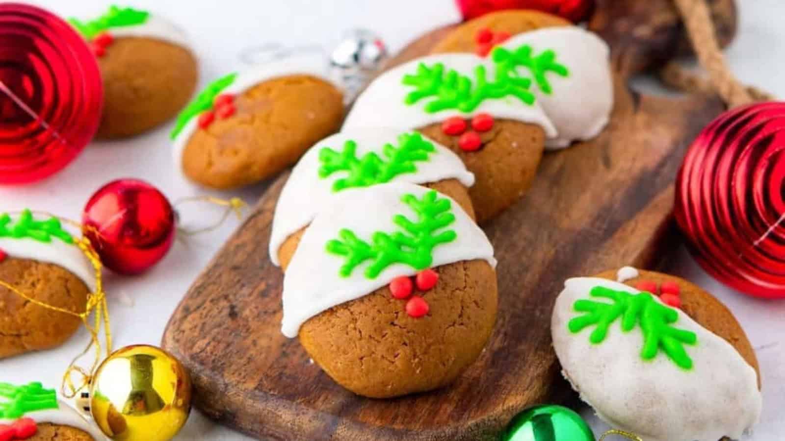 Festive cookies with white icing and green holly leaf designs topped with red berries, arranged on a wooden board. Surrounding the cookies are red, green, and gold Christmas ornaments.
