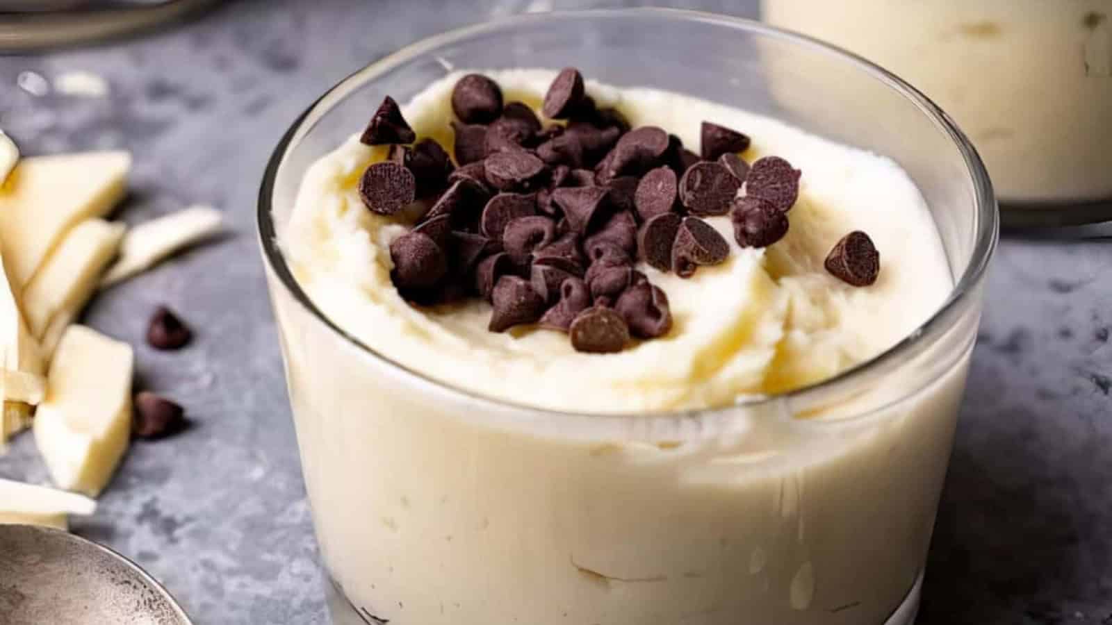 A glass bowl filled with creamy white dessert, topped with a generous amount of dark chocolate chips. The background shows a blurred surface with scattered chocolate chips and pieces of white chocolate.