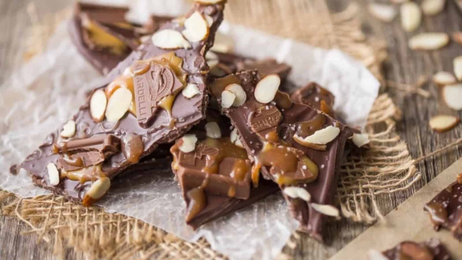 Pieces of chocolate bark topped with caramel drizzle and almond slices are placed on a piece of parchment paper, resting on a textured burlap surface.