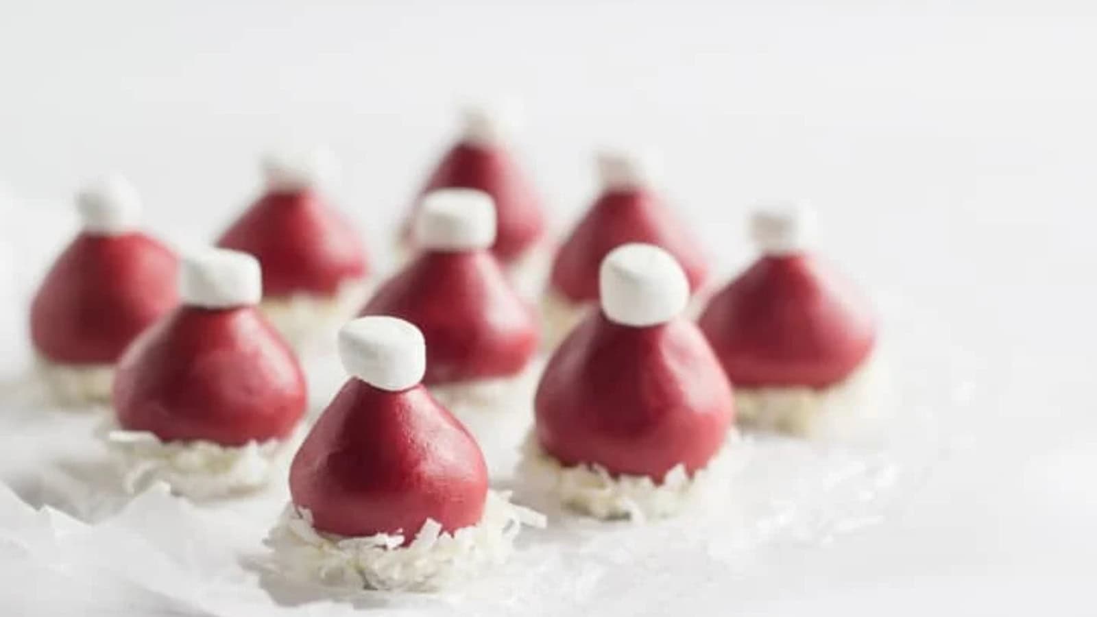 Nine red Santa hat treats made from strawberries, each topped with a small white marshmallow and resting on a bed of shredded coconut, arranged on a white surface.