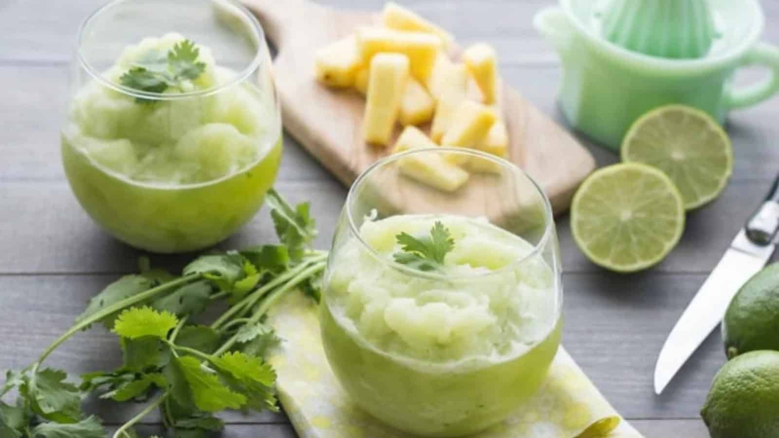Two glasses of a green slushy drink garnished with cilantro are on a table. Nearby are lime halves, a knife, cilantro sprigs, and pineapple pieces on a cutting board. A green citrus juicer is in the background.