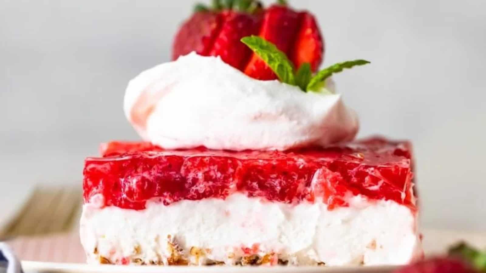 A slice of layered strawberry dessert on a plate. The bottom layer is crumbly, topped with a creamy white middle layer, a layer of red strawberry jelly, and garnished with whipped cream, a strawberry, and a mint leaf.