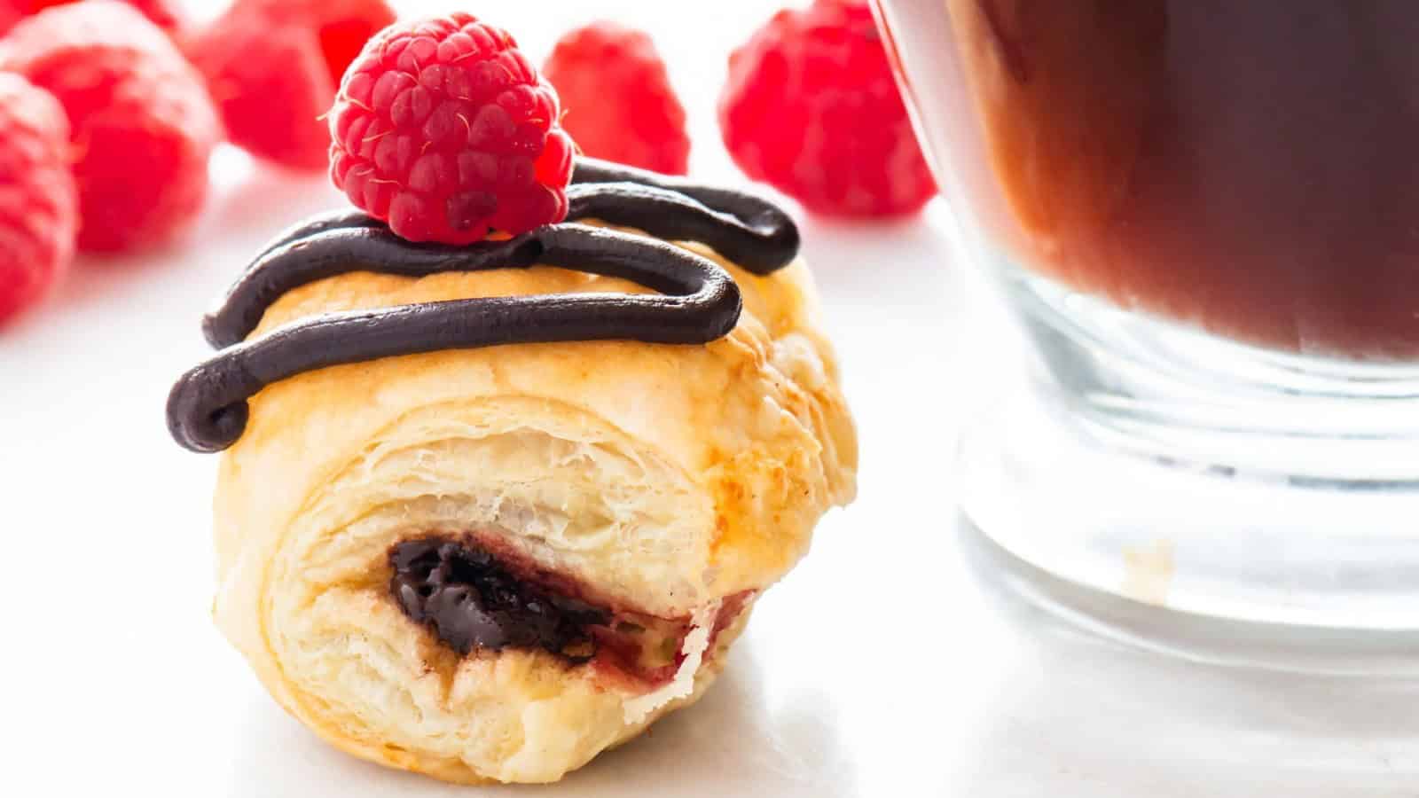 A flaky pastry topped with a fresh raspberry and chocolate drizzle rests near a glass of dark coffee. More raspberries are scattered in the background on a white surface.