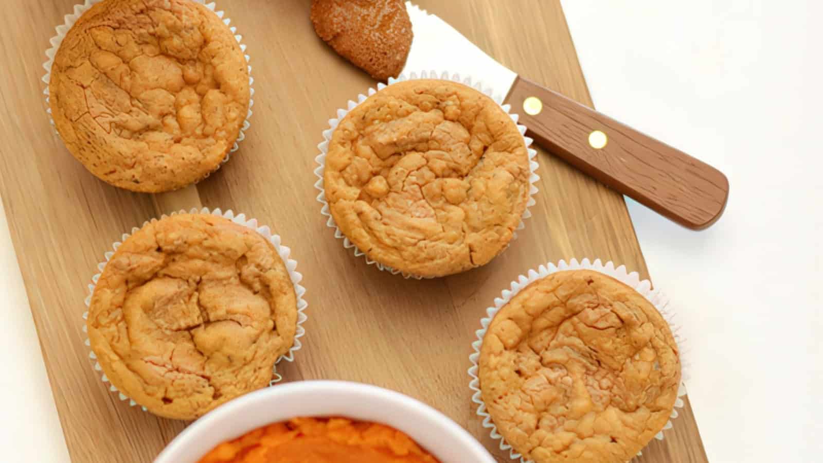 Four freshly baked muffins sit on a wooden board next to a knife with a wooden handle. There is a small amount of spread on the board. A bowl containing an orange spread, likely pumpkin or sweet potato, is partially visible.