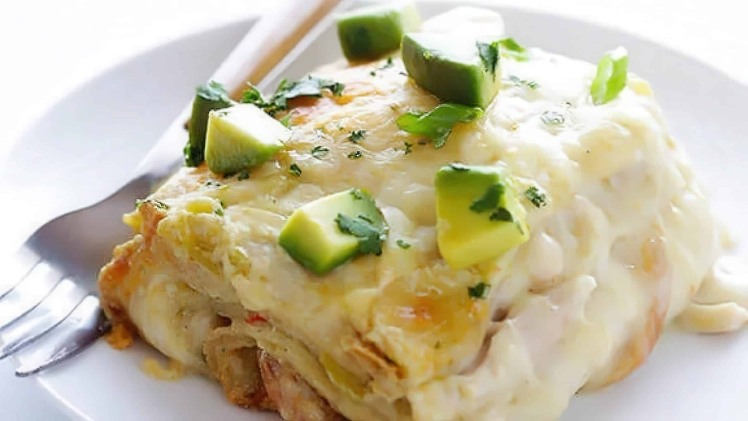 A slice of creamy enchilada casserole topped with diced avocado and cilantro, served on a white plate with a fork beside it.