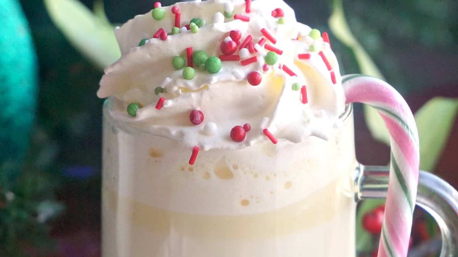 A mug filled with white chocolate is topped with whipped cream and colorful sprinkles, with a candy cane placed on the handle.