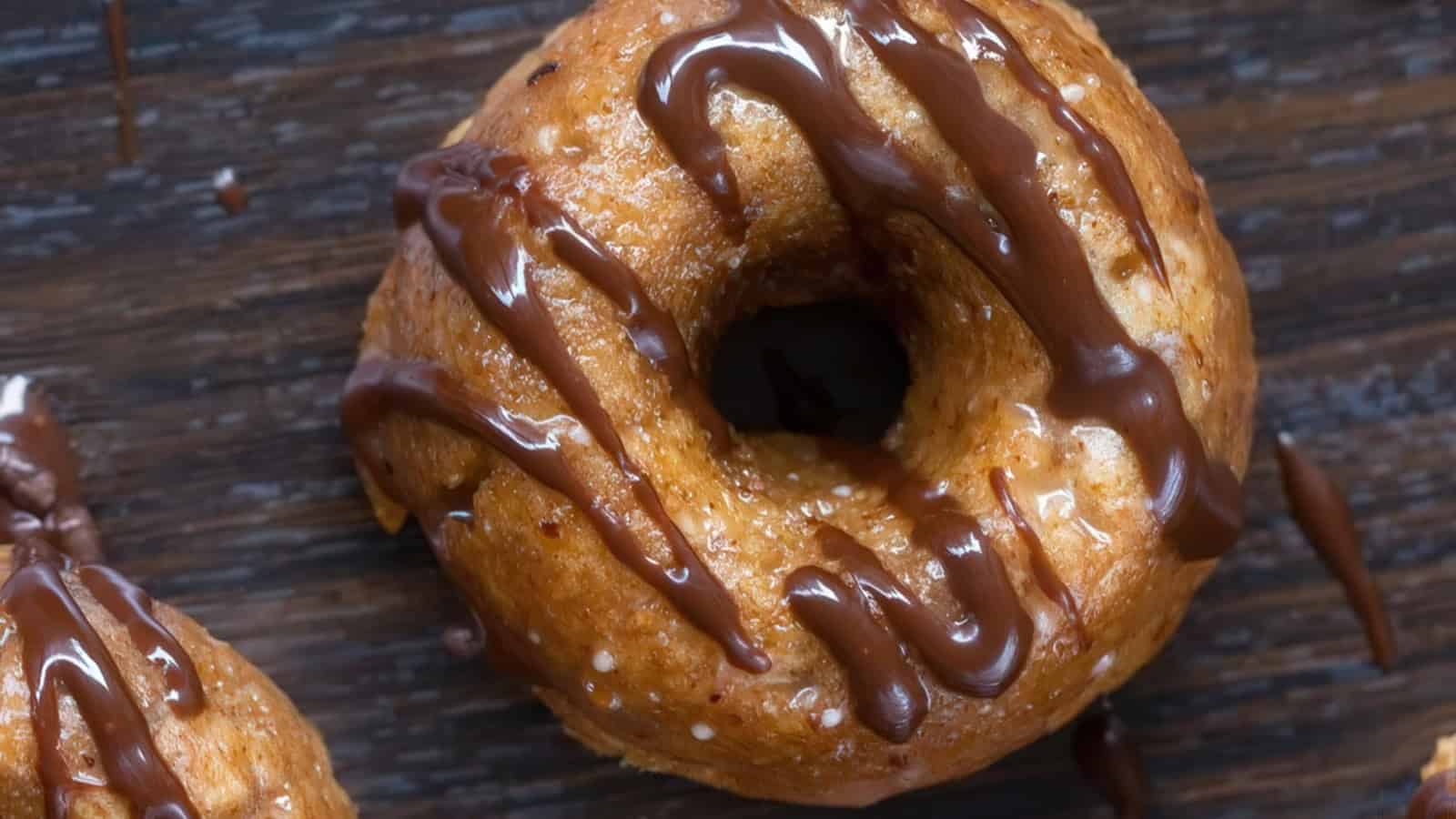 A close-up of a glazed doughnut topped with chocolate drizzle on a dark wooden surface.