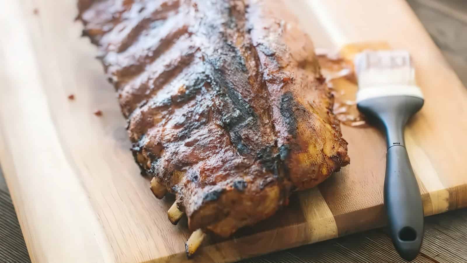 A rack of grilled ribs on a wooden cutting board, partially coated with barbecue sauce. A basting brush with sauce residue is placed beside the ribs. The ribs have a smoky, glazed appearance.