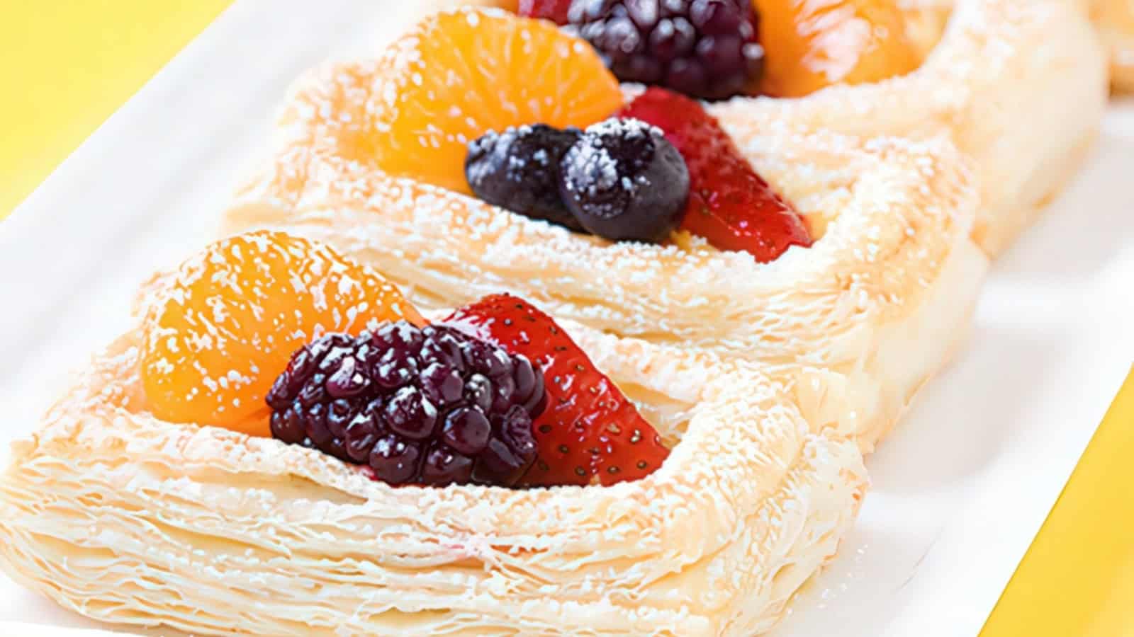 Flaky pastries topped with a mix of fresh fruit, including a blackberry, raspberry, mandarin slice, and strawberry. Dusting of powdered sugar is visible on the pastries, which are displayed on a rectangular white plate.