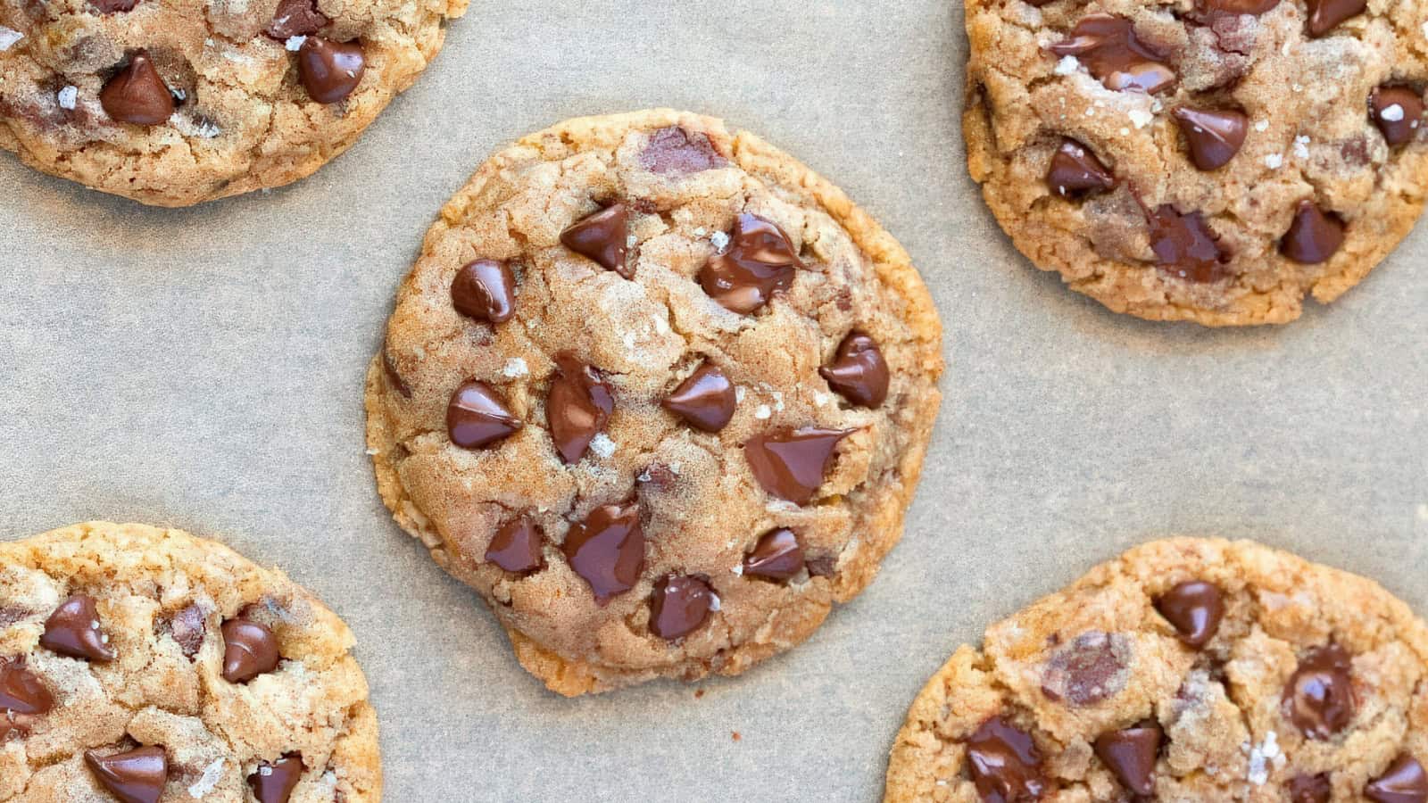 Close-up of five chocolate chip cookies on parchment paper. The cookies are golden brown with melted chocolate chips, showcasing a soft and chewy texture.