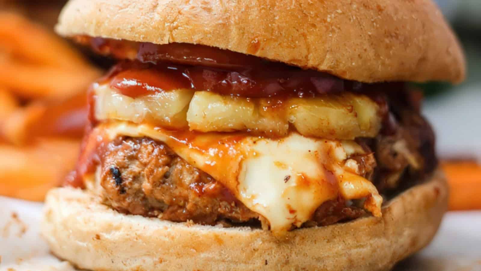Close-up of a burger with a grilled patty, melted cheese, pineapple slices, and BBQ sauce in a toasted bun. Sweet potato fries are blurred in the background.