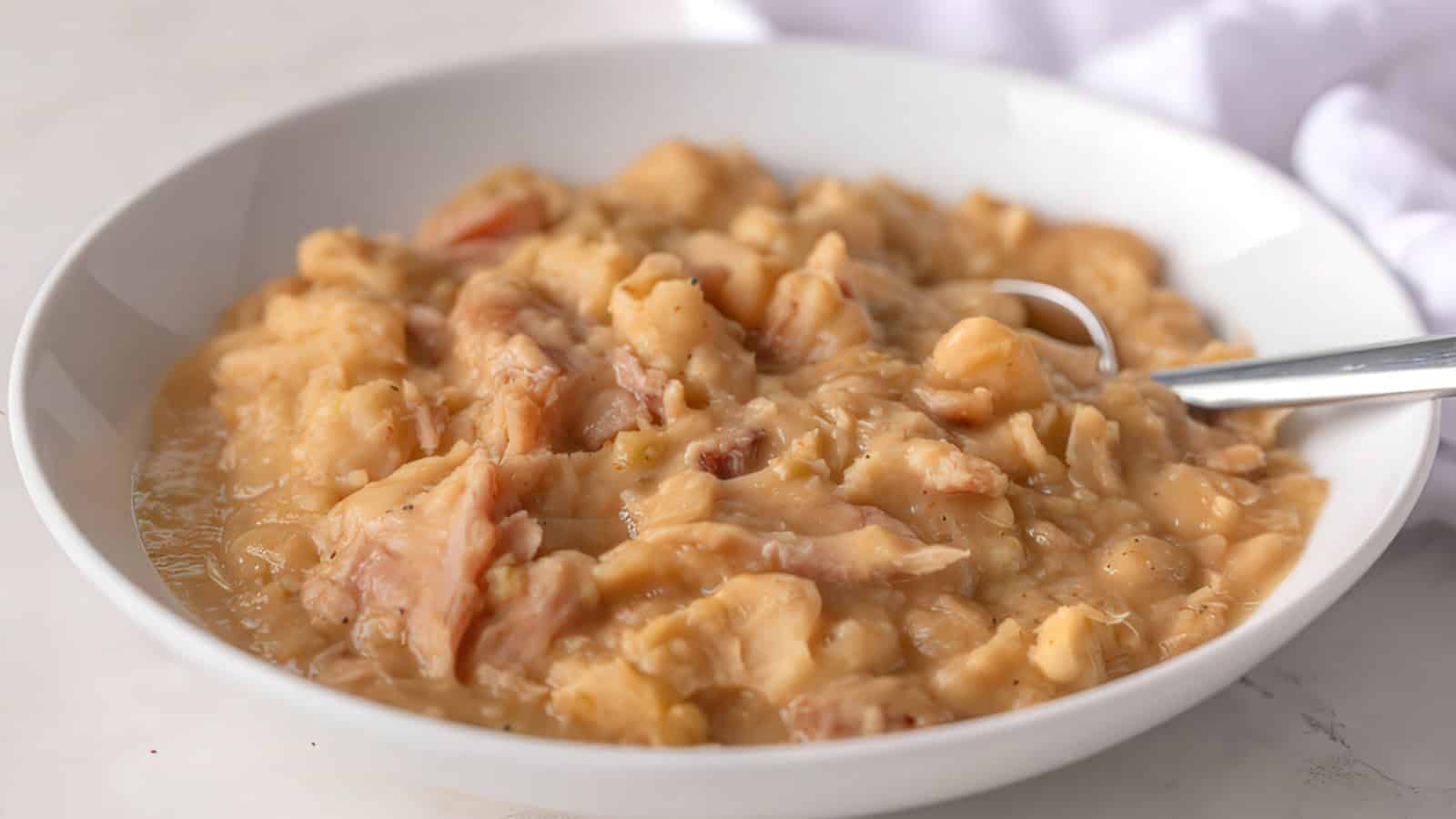 A bowl of creamy mashed potatoes with chunks, accompanied by a spoon, on a white surface.