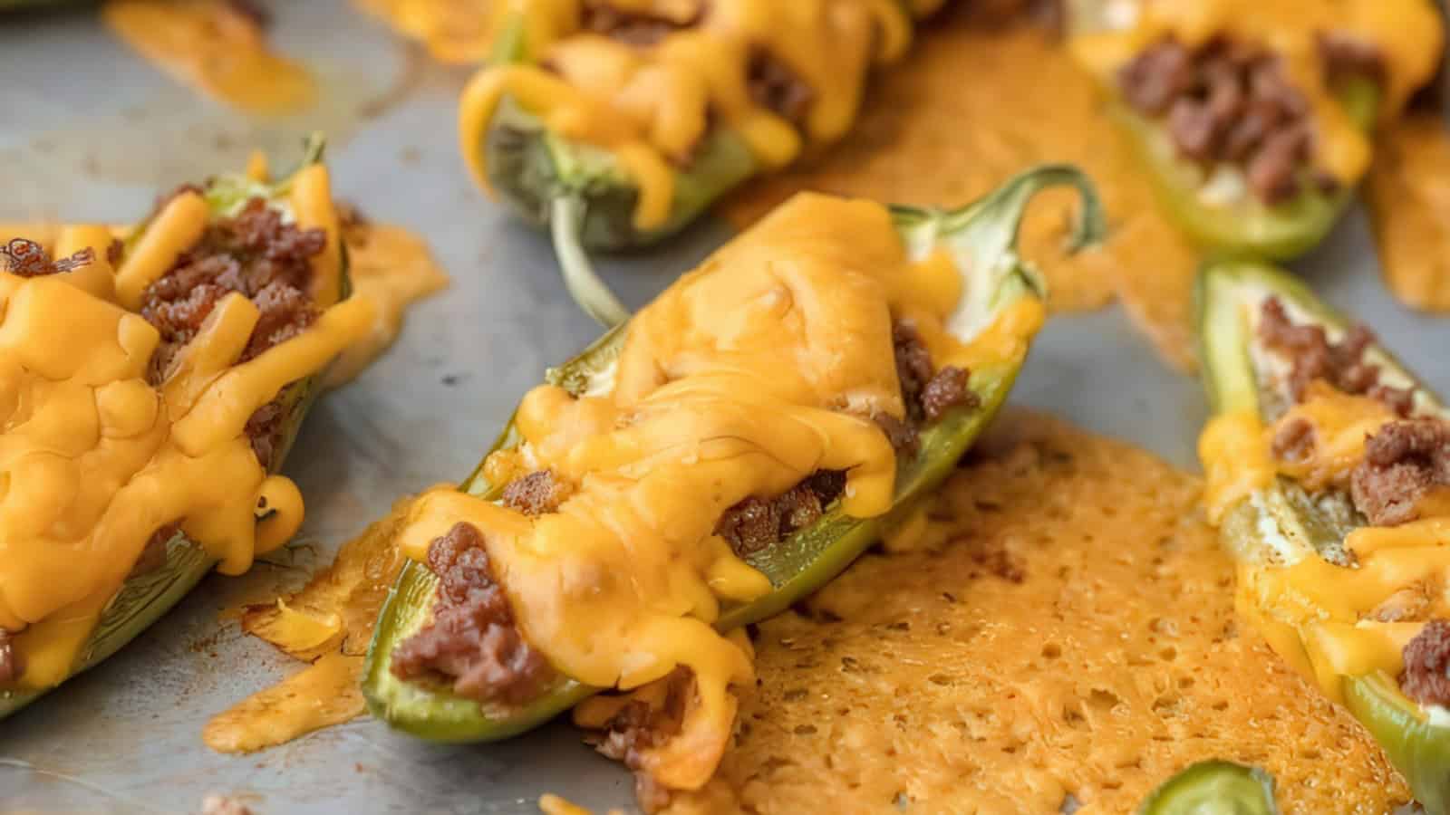 Stuffed jalapeño peppers topped with melted cheese are arranged on a baking sheet. The filling appears to be a mix of meat and cheese. A few jalapeños have slightly melted cheese spreading onto the sheet.