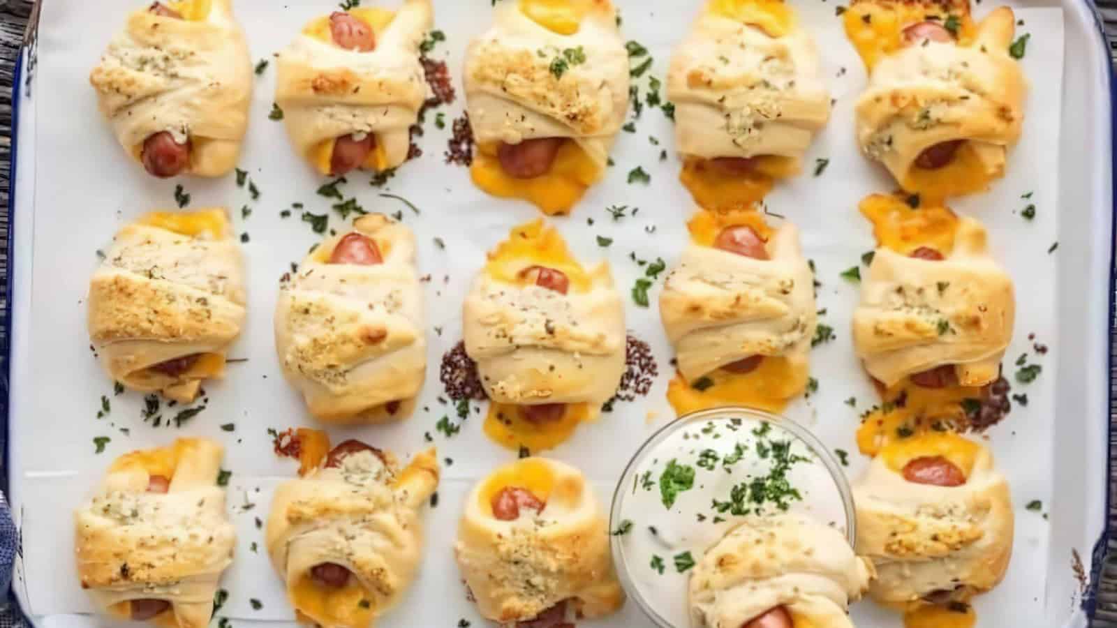Baked pigs in a blanket arranged on a tray, featuring small sausages wrapped in crescent dough with melted cheese. A small dish of creamy dipping sauce is in the center, garnished with chopped herbs.