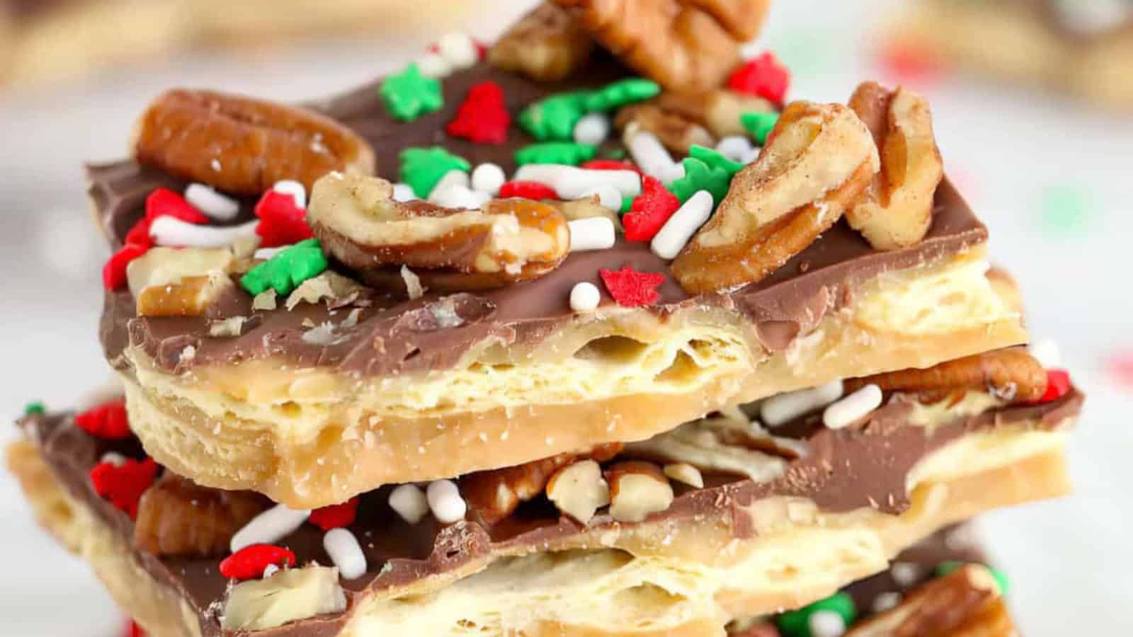 Close-up of stacked chocolate toffee bark topped with pecans, red and green star-shaped sprinkles, and white sugar sprinkles. The layers reveal a mix of chocolate and toffee on a crispy base.