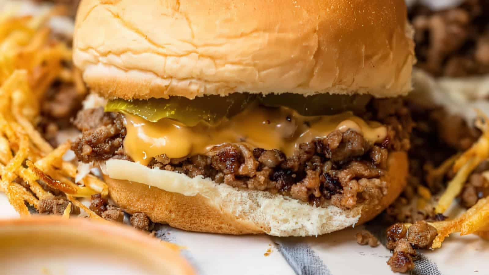 A juicy burger topped with melted cheese and pickle slices nestled in a soft bun. Crumbled beef and crispy fried onions are scattered around the burger, enhancing its savory presentation.