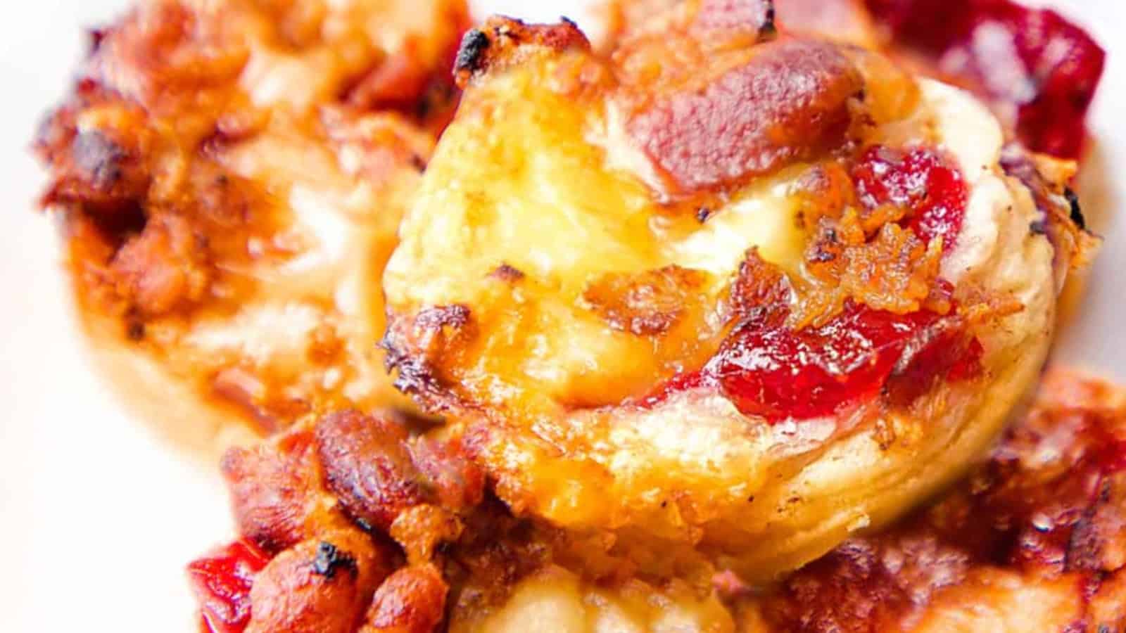 Close-up of a savory appetizer featuring bite-sized pastry cups filled with melted cheese, crispy bacon, and a topping of cranberry sauce. The golden-brown edges add a touch of crispiness to the dish.