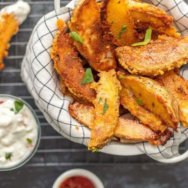 Seasoned potato wedges in a basket on a checkered cloth, with creamy dip and ketchup in small bowls next to them.