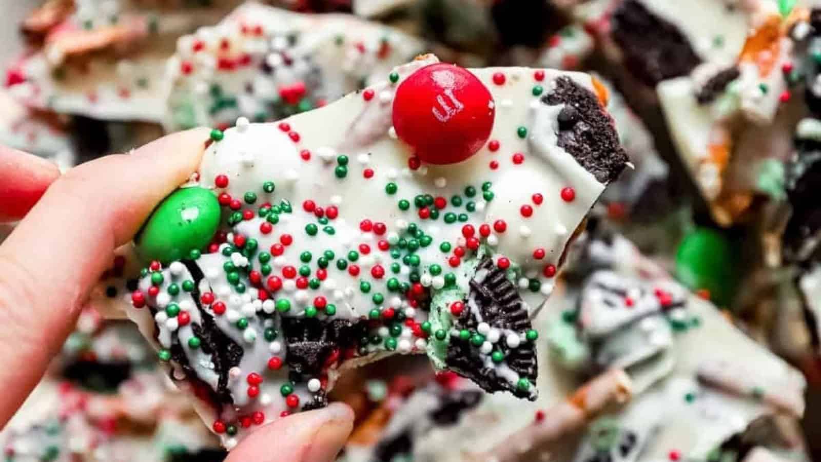 A hand holds a piece of festive bark candy covered in white chocolate, red and green sprinkles, and red and green candy-coated chocolates. Pieces of cookies and pretzels are mixed in, creating a colorful and textured holiday treat.