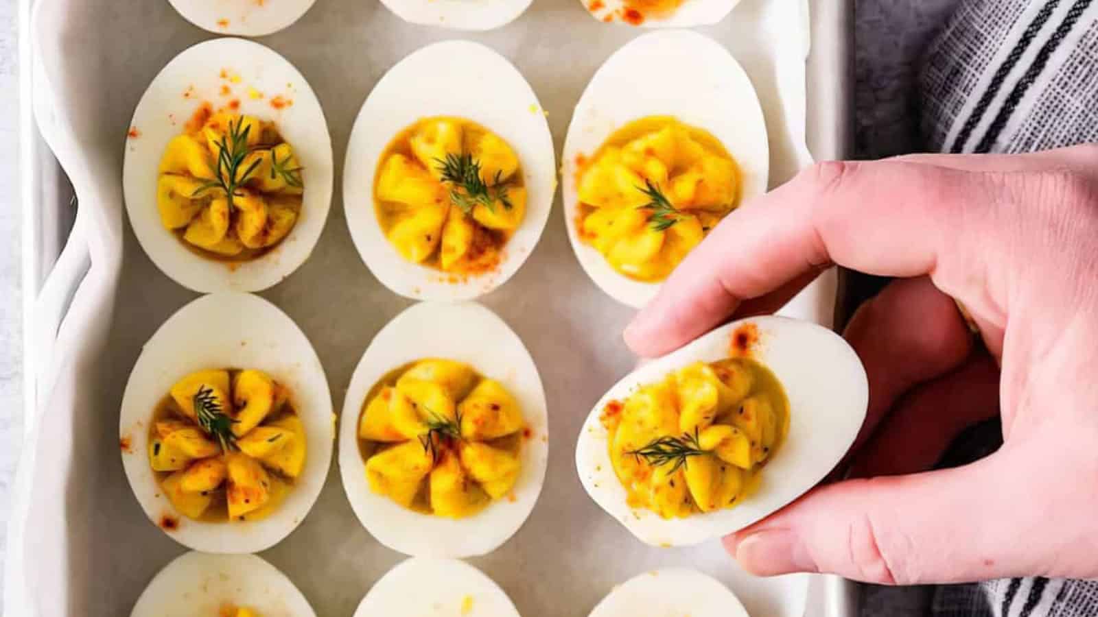 A hand holding a deviled egg over a tray filled with several deviled eggs. Each egg is garnished with dill and a sprinkle of paprika. The eggs are neatly arranged on a white dish with a striped cloth partially visible on the side.