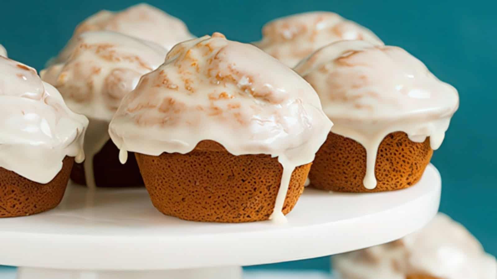 Close-up of six glazed muffins on a white cake stand against a blue background. The muffins are generously covered with a smooth, white icing that drips slightly down the sides.