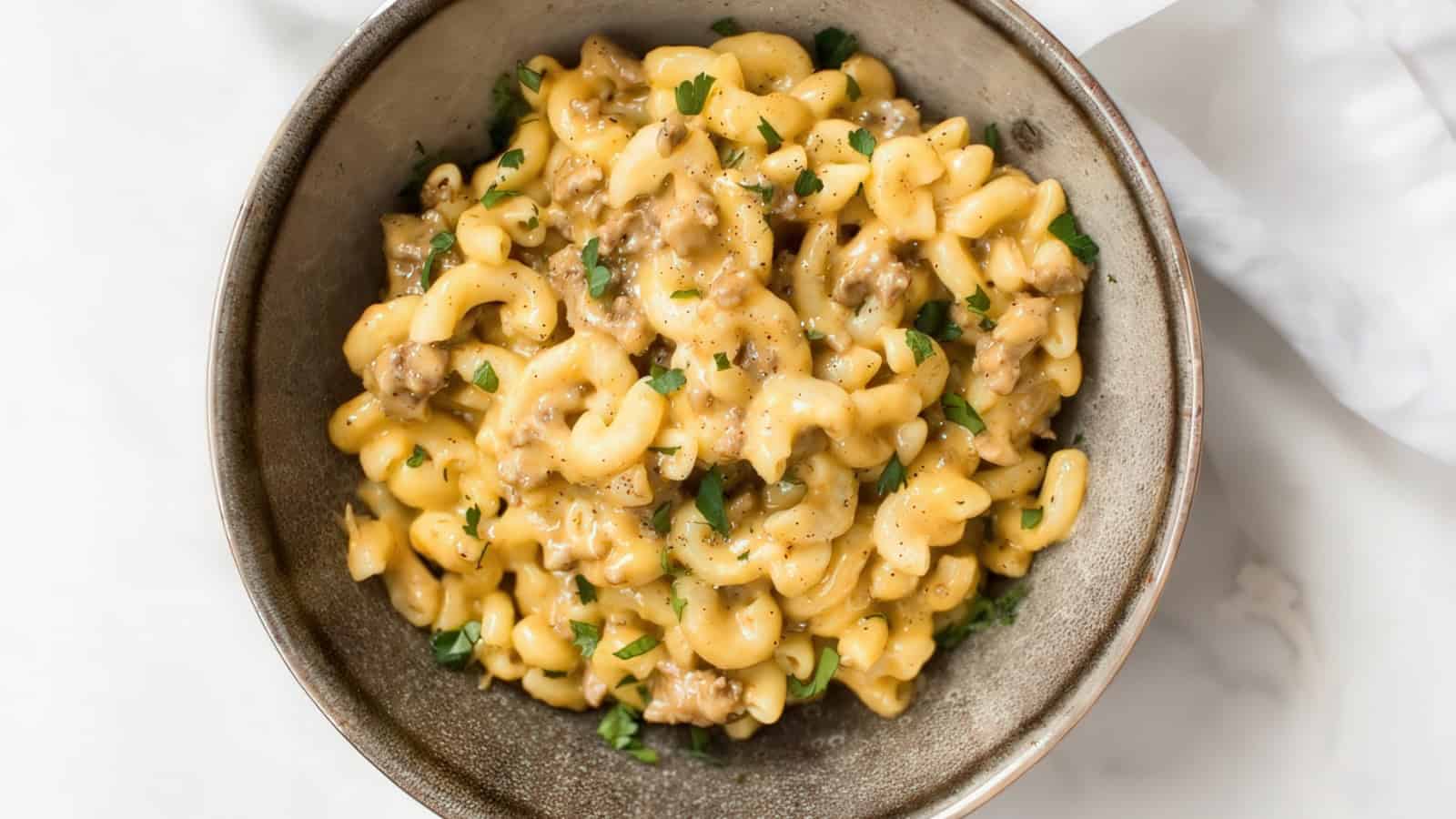 A bowl of creamy macaroni and cheese with ground beef garnished with freshly chopped parsley on top. The dish is served in a rustic, gray ceramic bowl on a white surface.