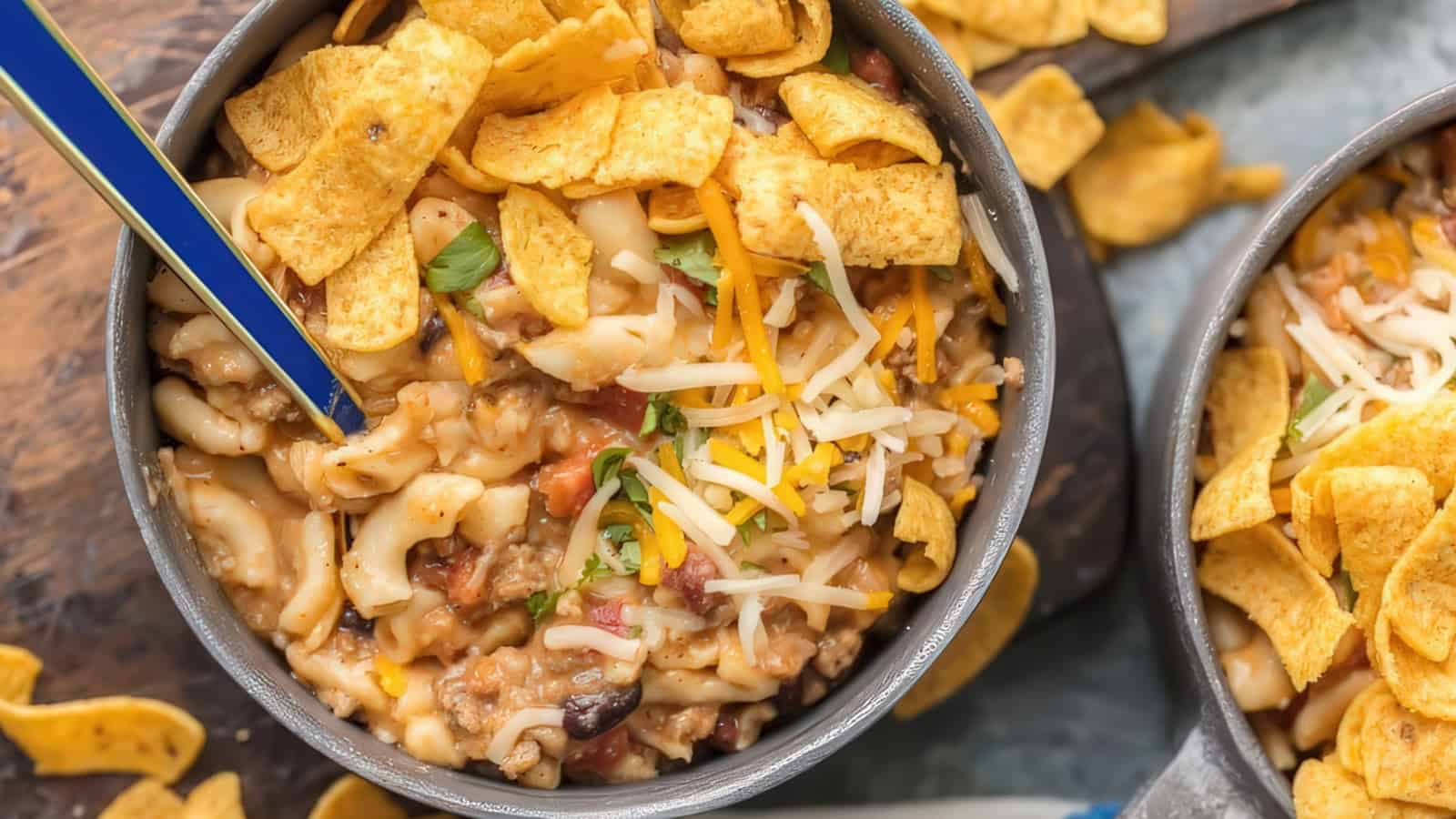 A bowl of cheesy chili macaroni topped with shredded cheese and corn chips. More corn chips are scattered around the bowl on a wooden surface. The dish is garnished with herbs.