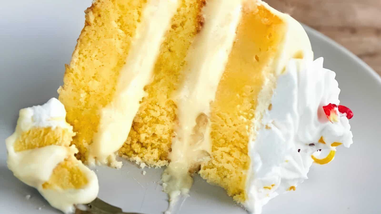 A slice of yellow cake with creamy frosting between the layers and a dollop of whipped cream on the side, served on a white plate. A fork with a small piece of cake rests beside the slice.