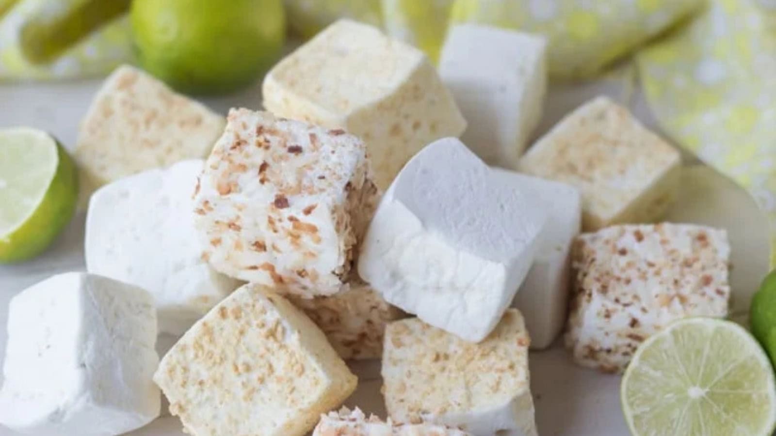 A pile of homemade marshmallows, some plain white and others sprinkled with toasted coconut, is arranged on a surface. Halved and whole limes are scattered around, accompanied by a yellow patterned cloth.
