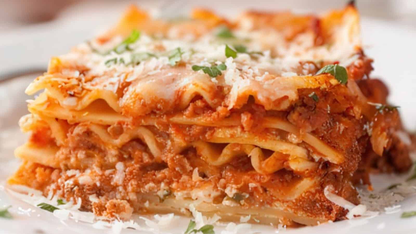 A close-up of a slice of lasagna on a plate, featuring layers of pasta, rich meat sauce, and melted cheese. The top is garnished with grated cheese and fresh herbs.