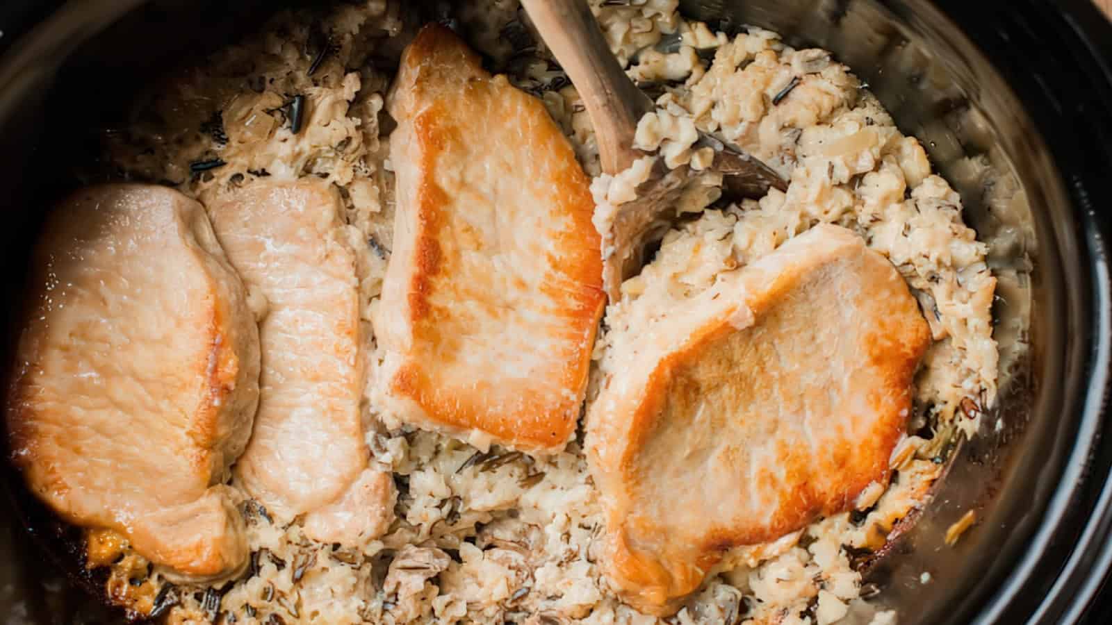Three cooked pork chops placed on top of a creamy mixture of rice in a slow cooker, with a wooden spoon partially submerged in the mixture on the right side. The scene has a warm, appetizing appearance.