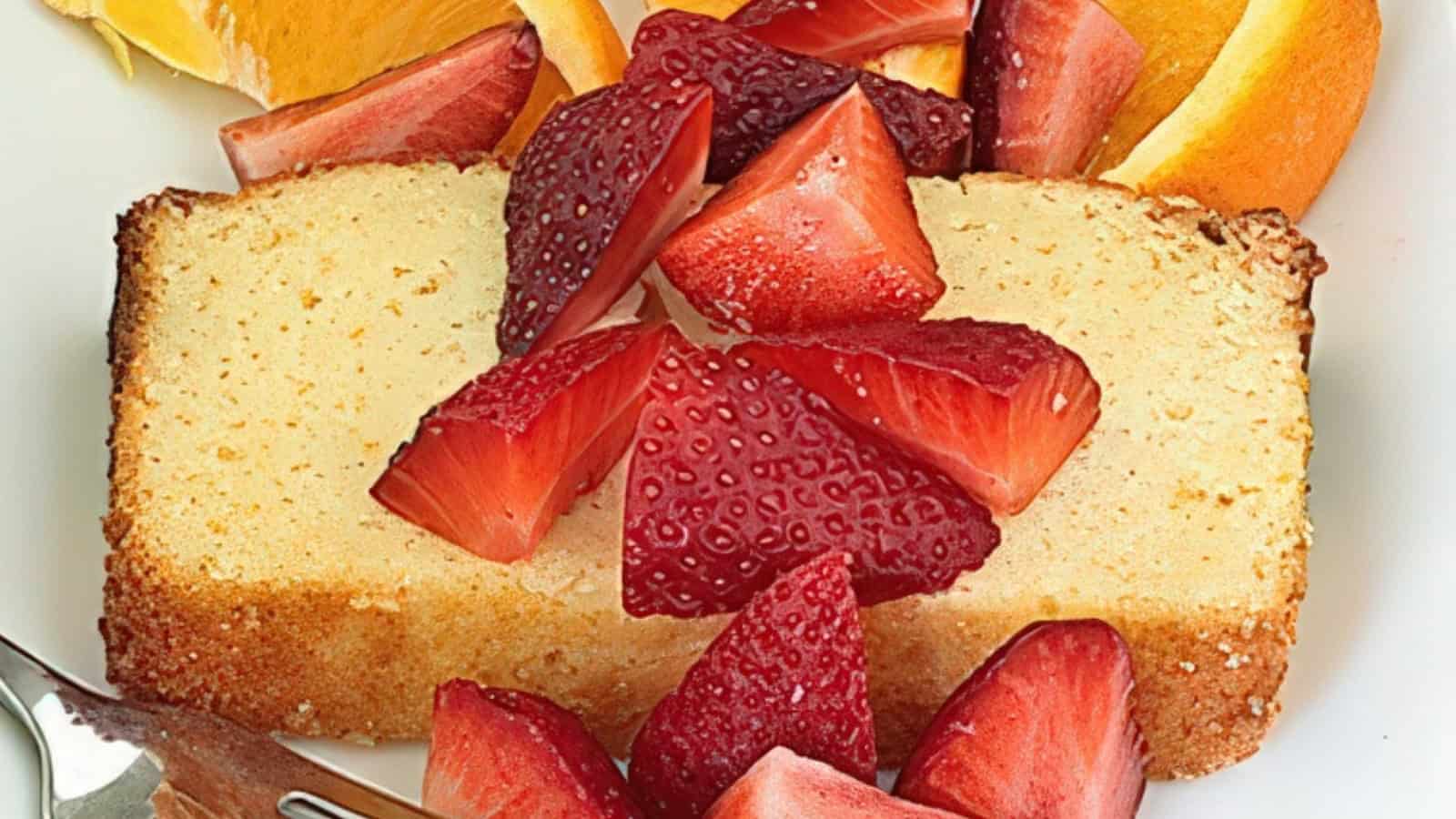 A slice of pound cake topped with fresh strawberries is placed on a plate alongside orange wedges. A fork is positioned nearby, ready for use.